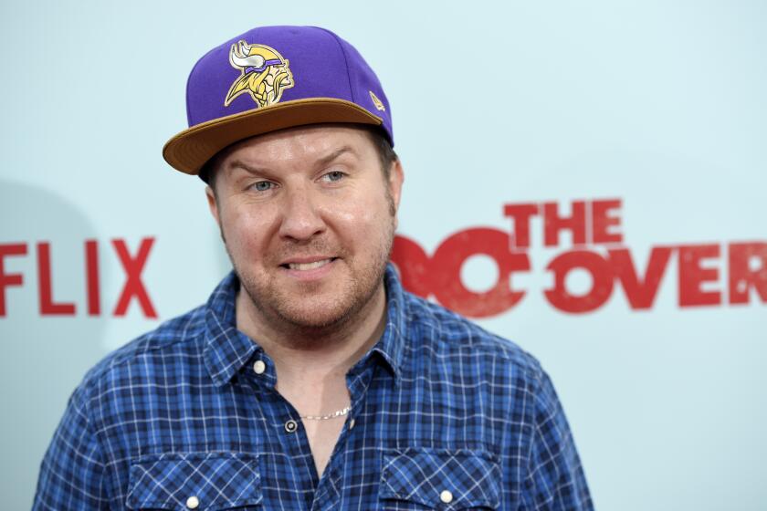 A white man with facial hair, a purple hat and a blue plaid shirt smiling and looking to his side in front of a blue backdrop