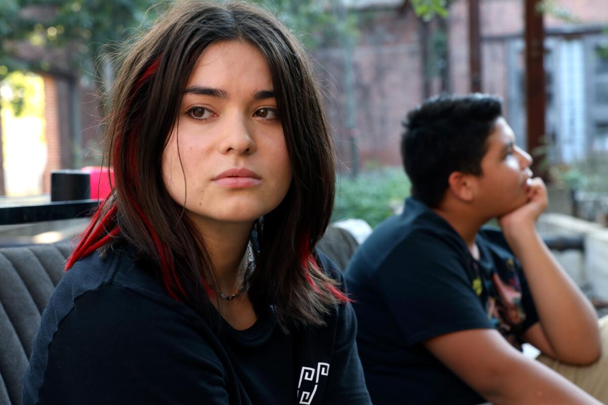 Una niña con cabello largo y castaño mira hacia un lado mientras está sentada cerca de un niño con la barbilla apoyada en la mano en un automóvil abierto.