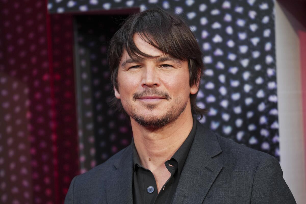 Josh Hartnett smiling slightly and squinting in a dark gray suit jacket and black open-collar shirt