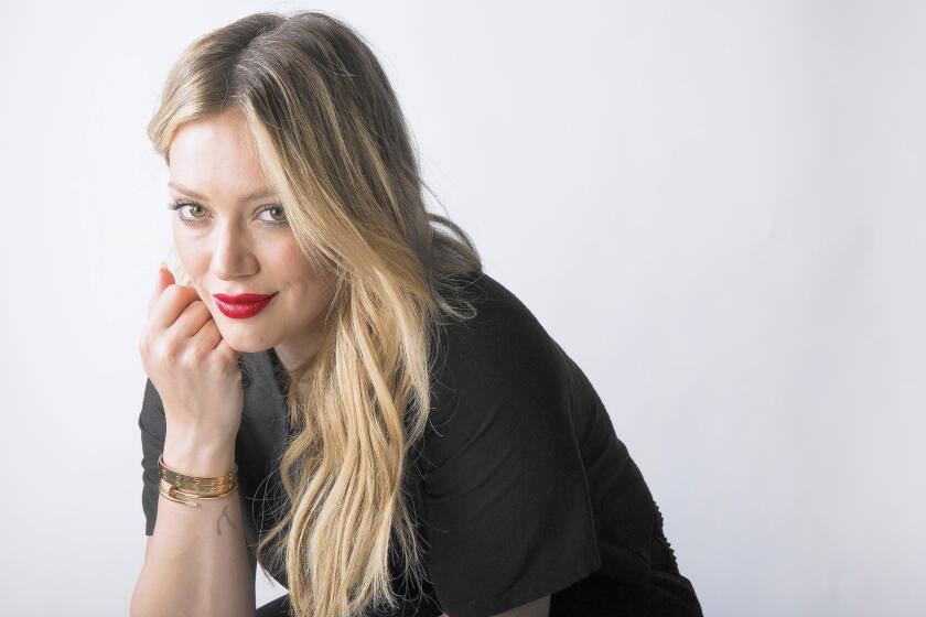 Hilary Duff in a black T-shirt and pants, sitting with her chin resting on her hand