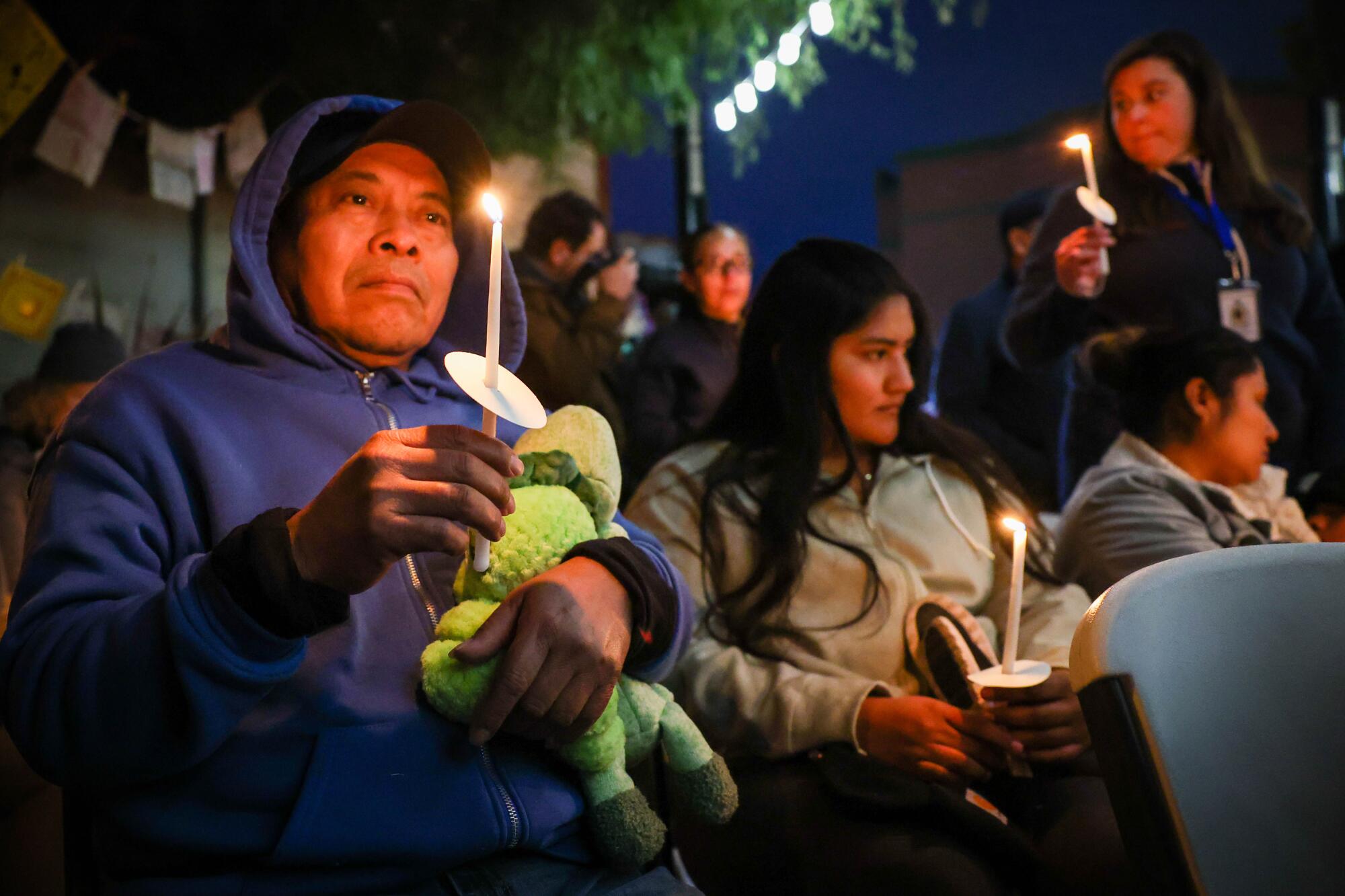 A man is holding a candle and stuffed animal and is sitting next to others at a vigil