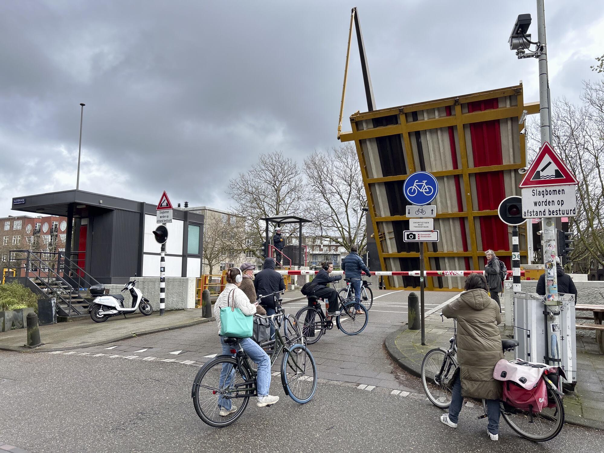 A moped parked outside a small, tidy building next to a raised drawbridge as several people on foot or bicycle wait to cross