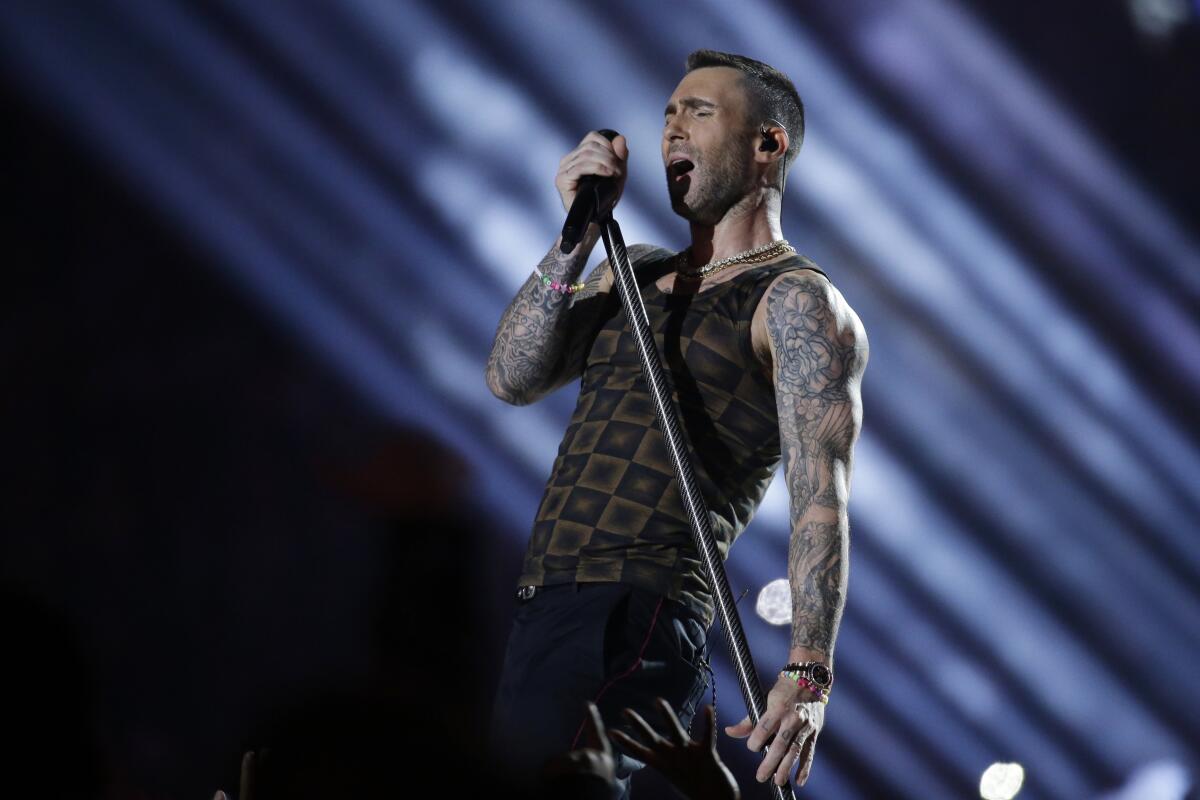 A man in a tank top with heavily tattooed arms sings into a microphone attached to a stand in concert