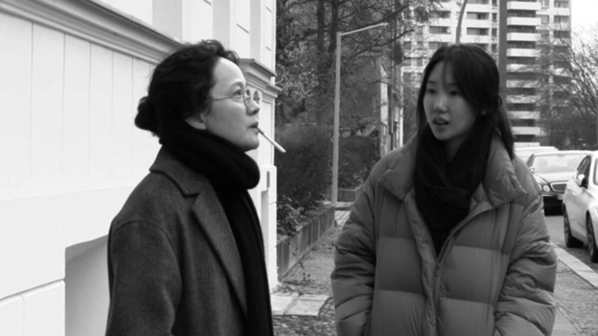 Two women stand outside, one of them smoking.