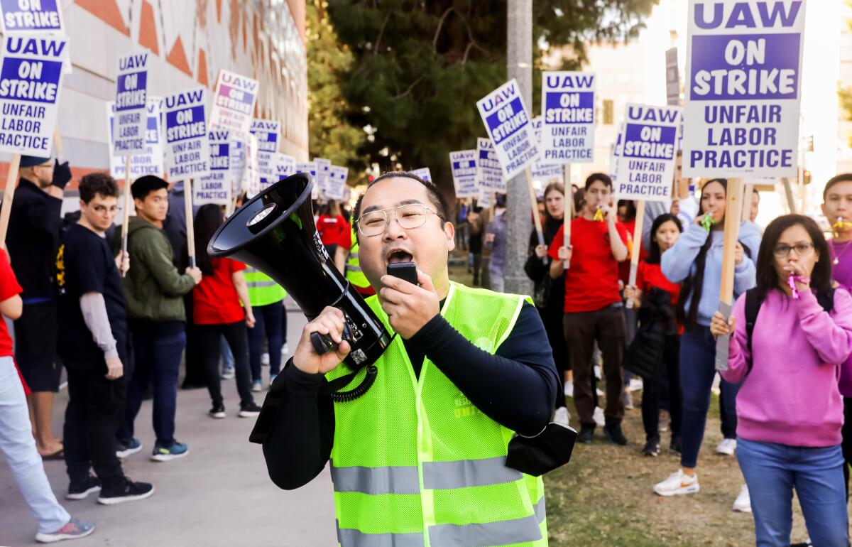 A man in a high-visibility vest talks into a megaphone as protesters hold signs that say UAW on strike, unfair labor practice