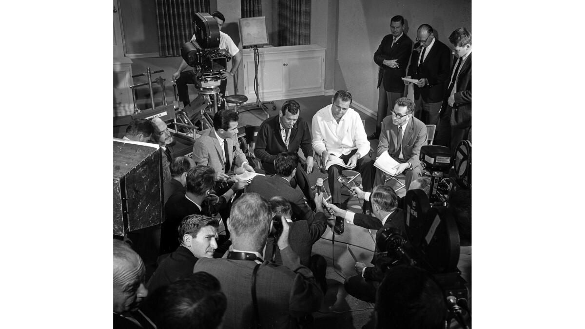 Four men sit in chairs, surrounded by men with cameras and microphones