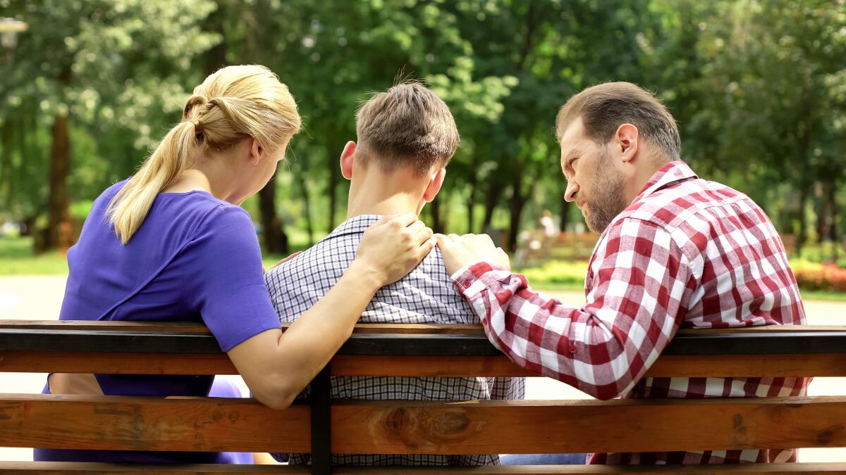 A woman with blond hair, in a purple top, and a man in a red plaid shirt flank a teenage boy on a bench