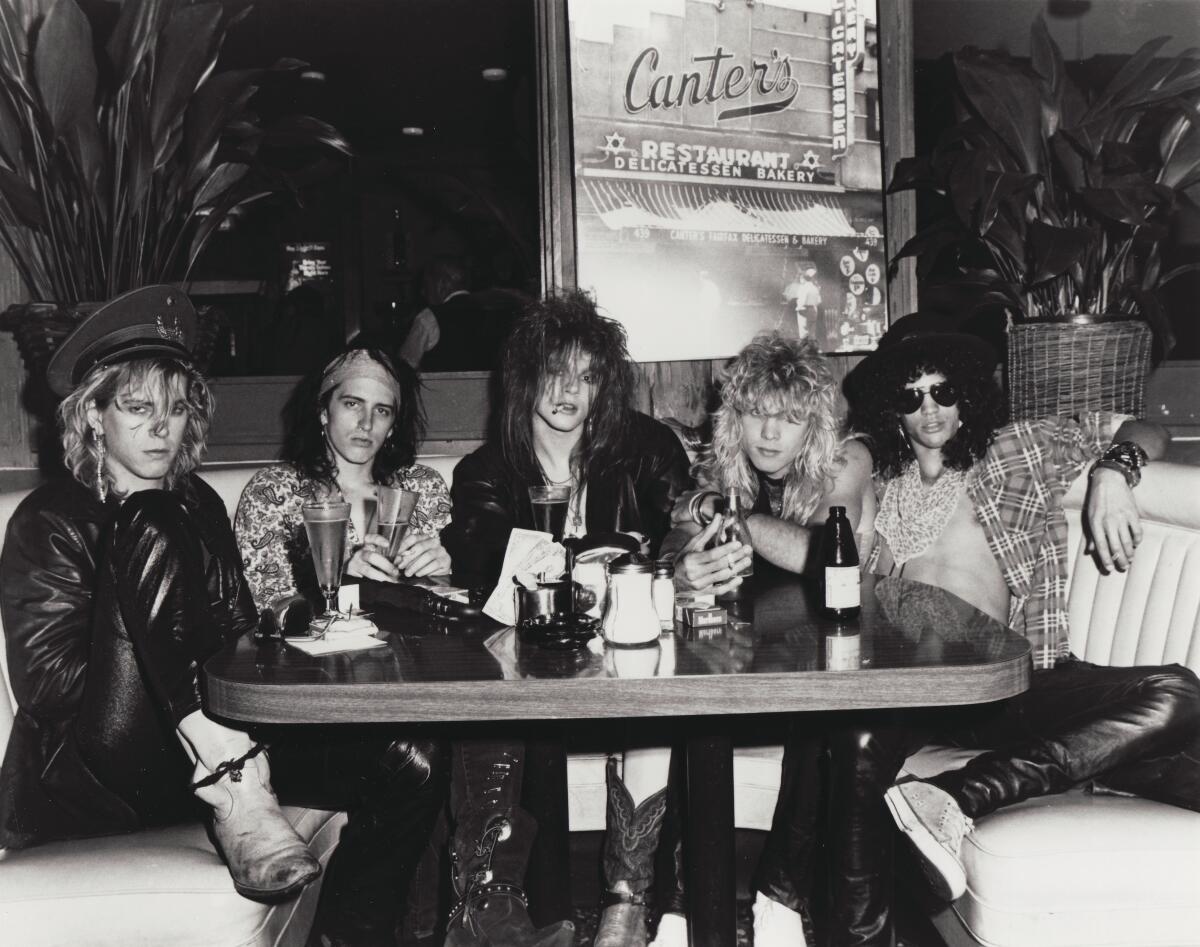 Five young men, two in leather pants and two in cowboy boots, with long, tousled hair sit in a booth at a deli.