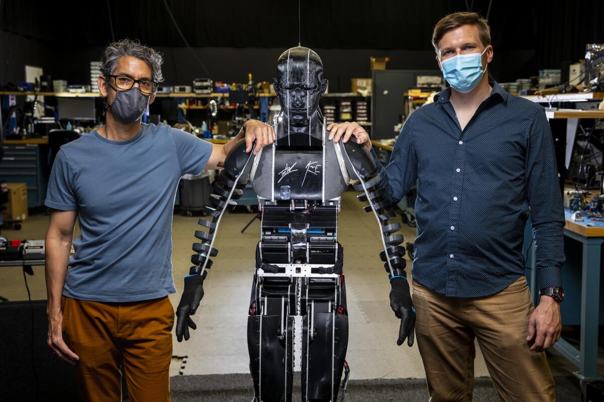 Two men resting their hands on the shoulders of a humanoid robot, center