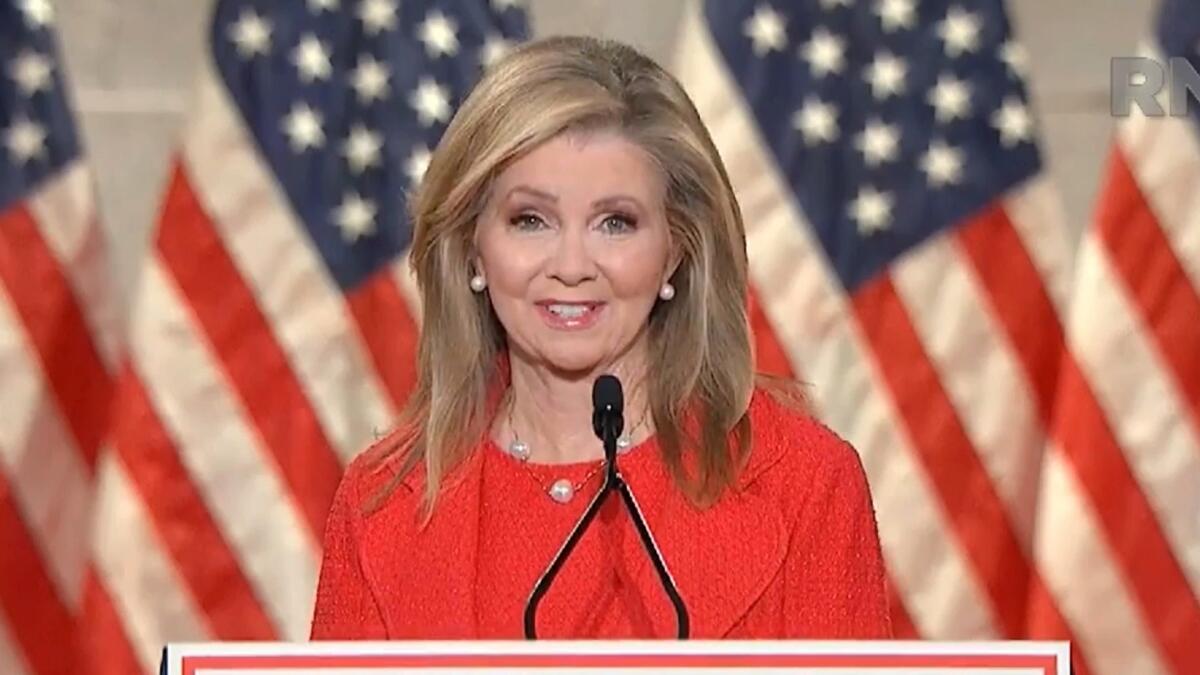A woman in red business attire at a podium in front of American flags