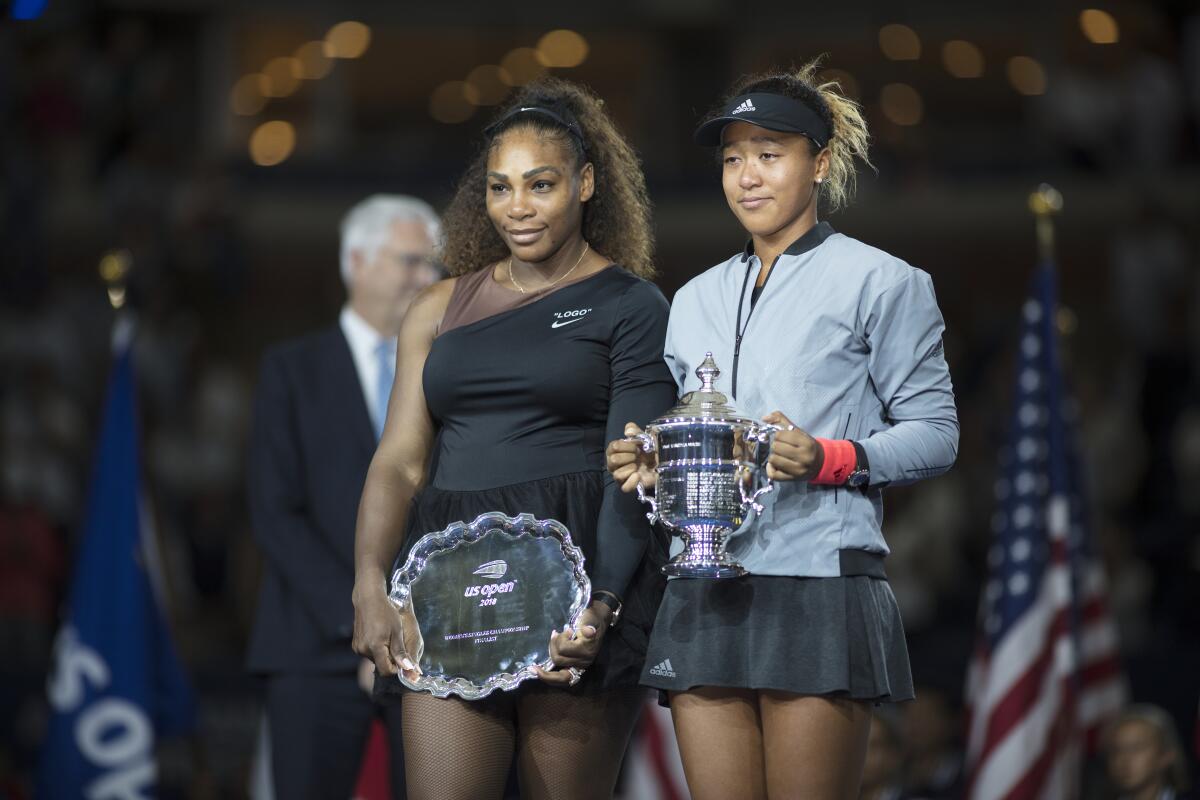 Two women in tennis uniforms stand side by side. The woman on the right holds a trophy, the other holds a silver platter.