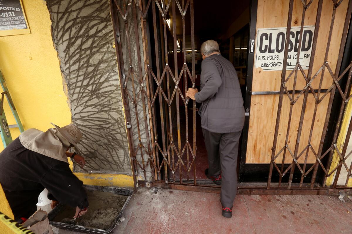 A man, seen from behind, opens a folding exterior metal door as he enters a business with a boarded-up front door.