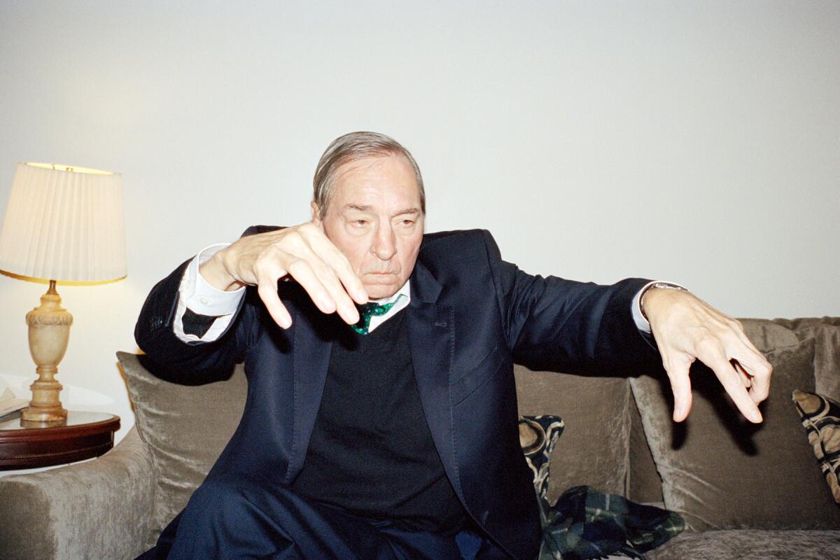 A flash-saturated photo shows William Eggleston sitting on a velvet couch while making a gesture of conducting music