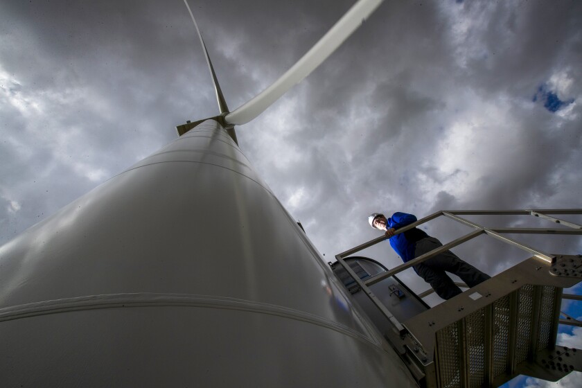 A man in a hard hat stands on a staircase next to a huge wind turbine, whose blade is seen above.