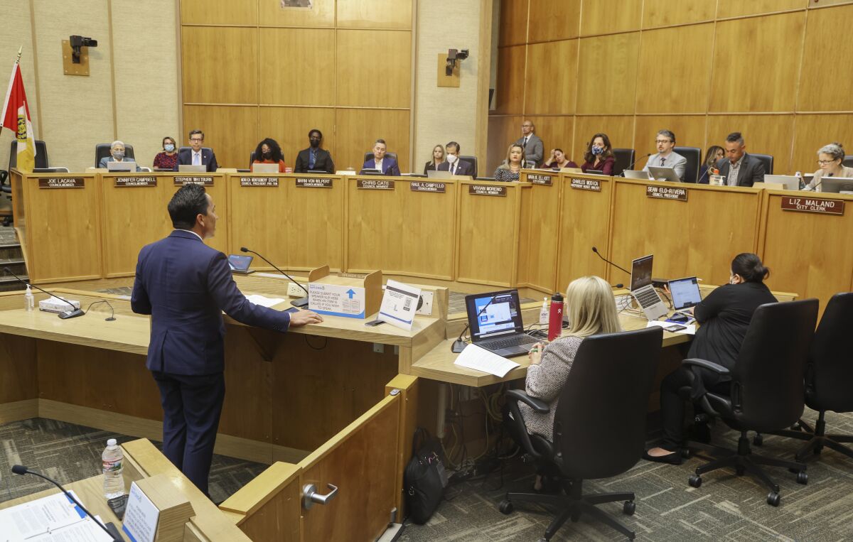 A man in a suit speaks at a podium in council chambers to a panel of arrayed council members.