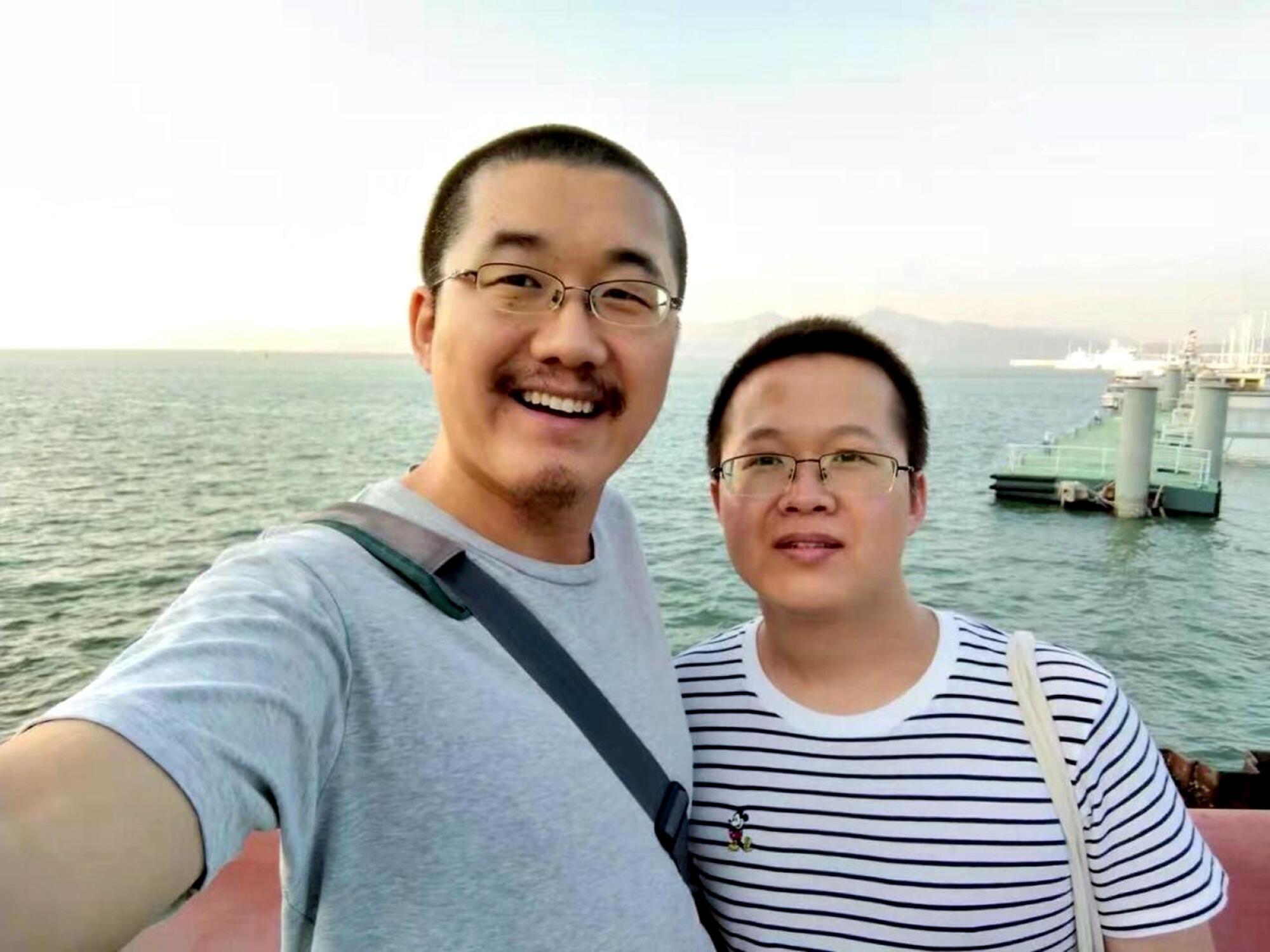 Two people in glasses pose for a photo with the ocean in the background
