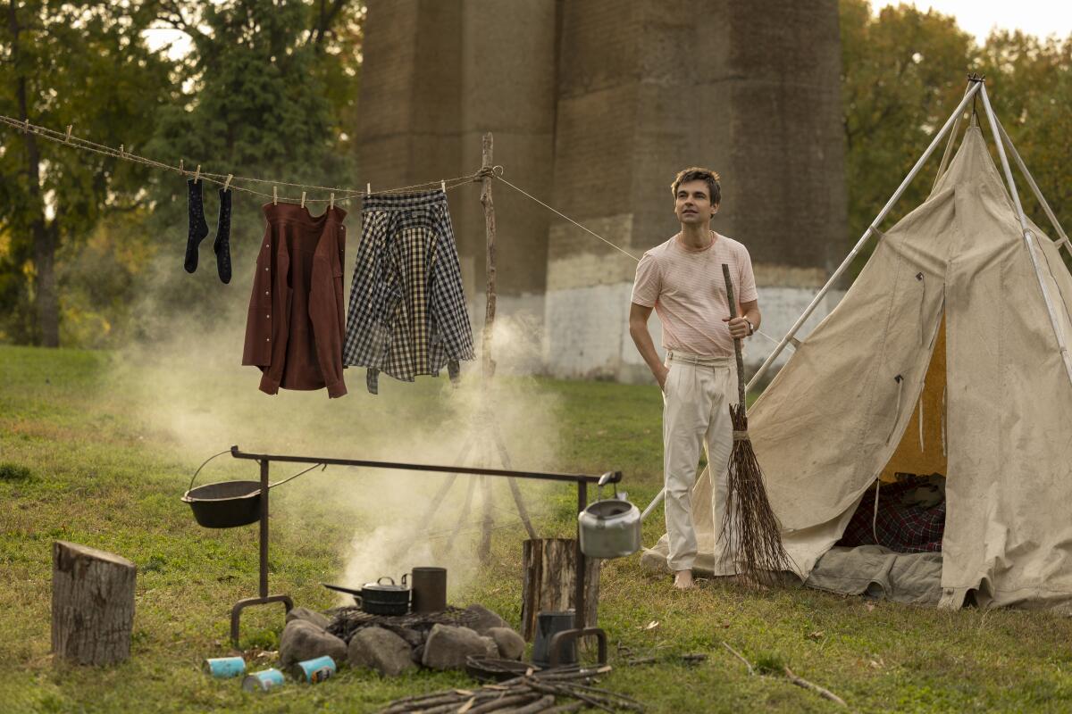 A man stands between a clothesline and a cloth tent that is in front of a a campfire.