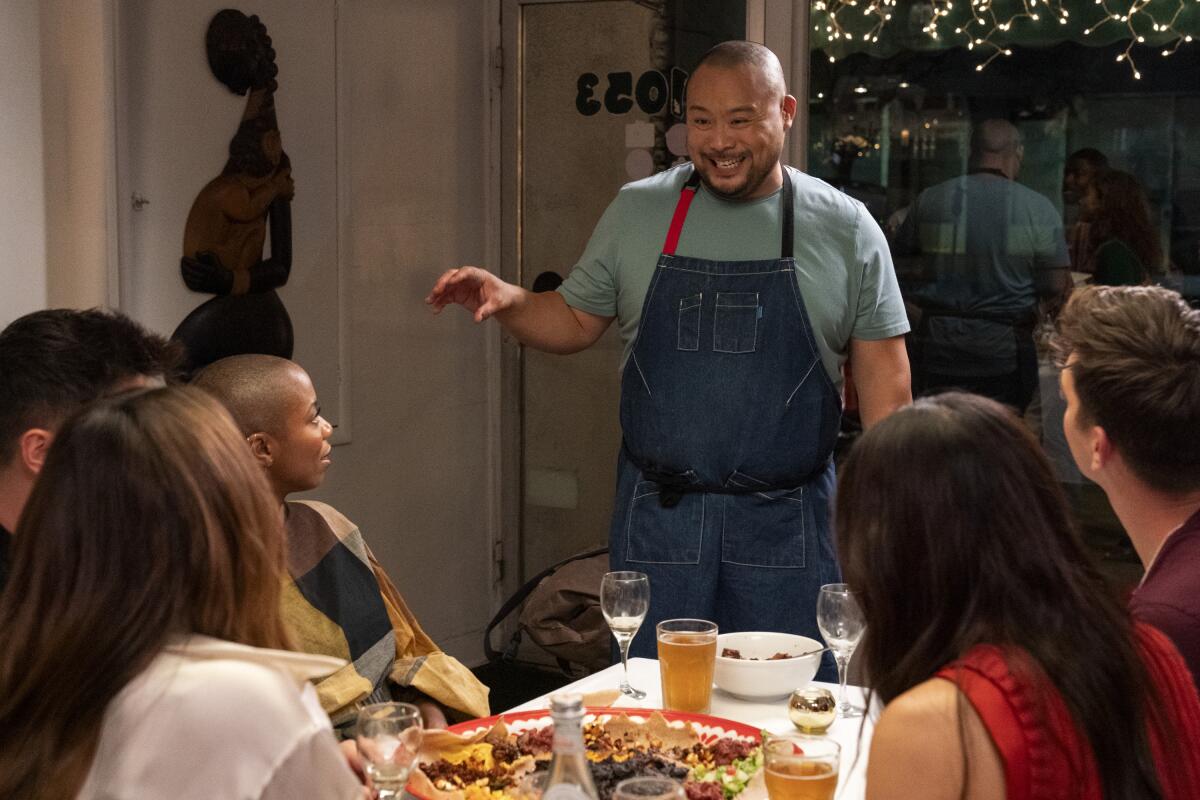 David Chang in a blue apron stands at the head of a dining table.