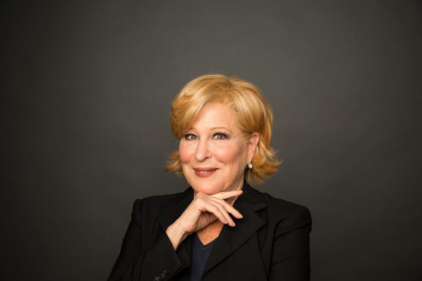 Bette Midler poses with hand under her chin, in black suit before dark studio background