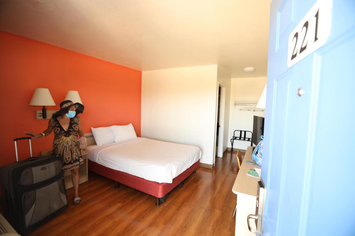 A woman stands next to a bed in a hotel room with her rolling luggage.