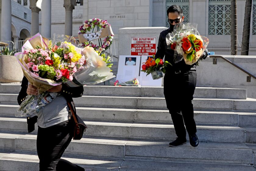 LOS ANGELES, CA - JANUARY 21: Marina Sastre, a court interpreter and friend of Daniel Felix, and Hector Quezada, fiance' of Masiel Felix, daughter of Daniel Felix, remove the flowers at end of a vigil held on the steps of City Hall in downtown on Thursday, Jan. 21, 2021 in Los Angeles, CA. A photo of Daniel Felix can be seen in the background. A vigil was held to honor Sergio Cafaro, 56, and Daniel Felix, 66, two court interpreters at Los Angeles Superior Court who died recently of COVID-19. (Gary Coronado / Los Angeles Times)