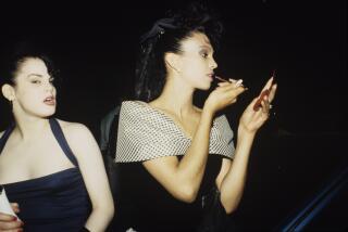 Carmen Xtravaganza stands next to another person, wearing a black outfit, staring into a compact mirror, applying makeup 