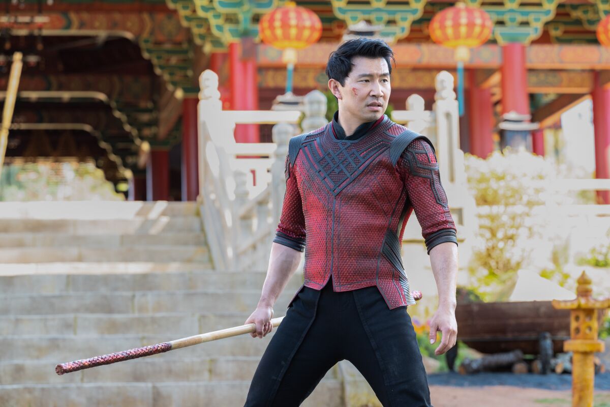 A man in a superhero costume stands before a stone temple's steps, holding a wooden pole in one hand.