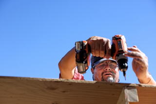 A man's head and hands against a clear blue sky as he holds a drill over a length of wood