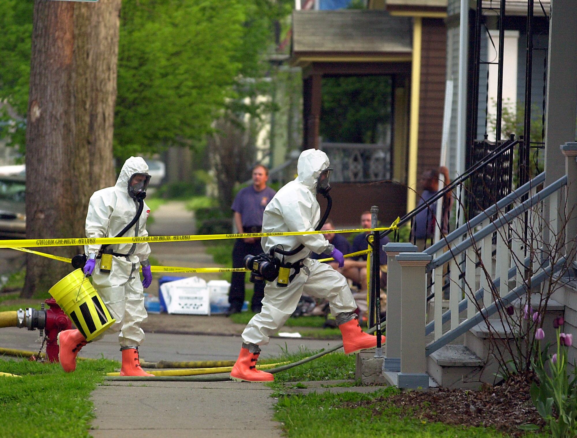 Two people in white hazmat suits and respirators head toward the steps of a home cordoned off with yellow tape