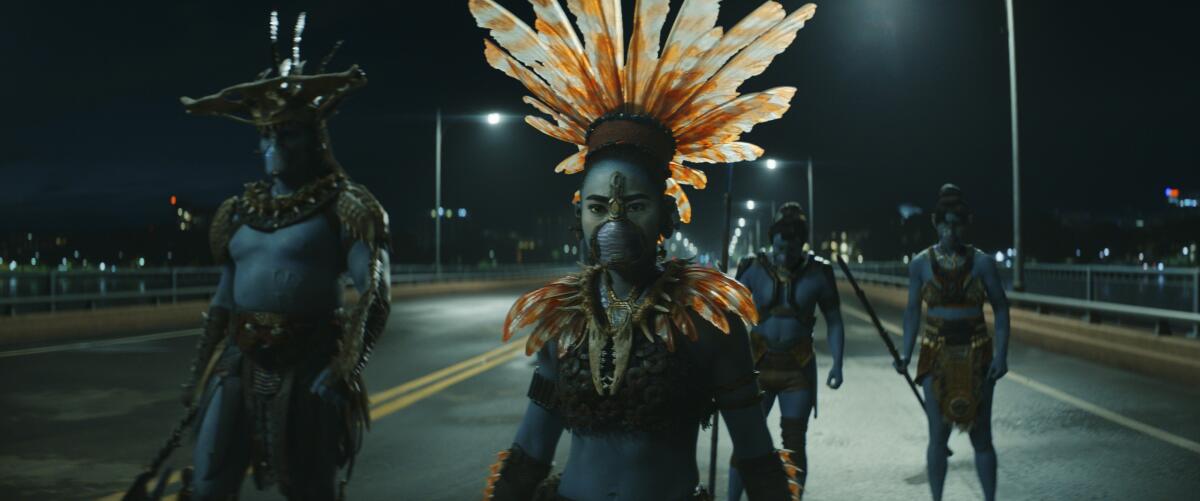A woman warrior stands with three male warriors dressed in costumes of beads and feathers in the "Black Panther" sequel.