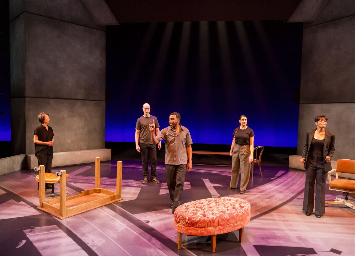 Five people stand on a stage amid upright and overturned furniture.