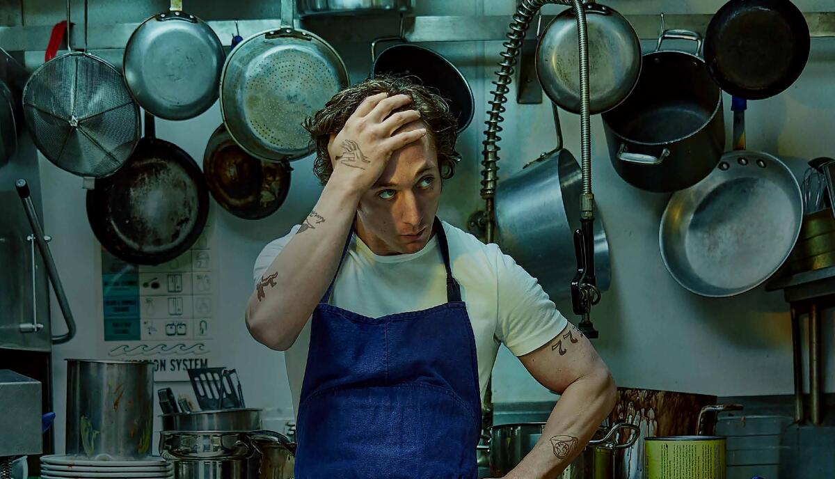 A chef stands in a kitchen, hand on forehead, with many metal pans hanging behind and above him
