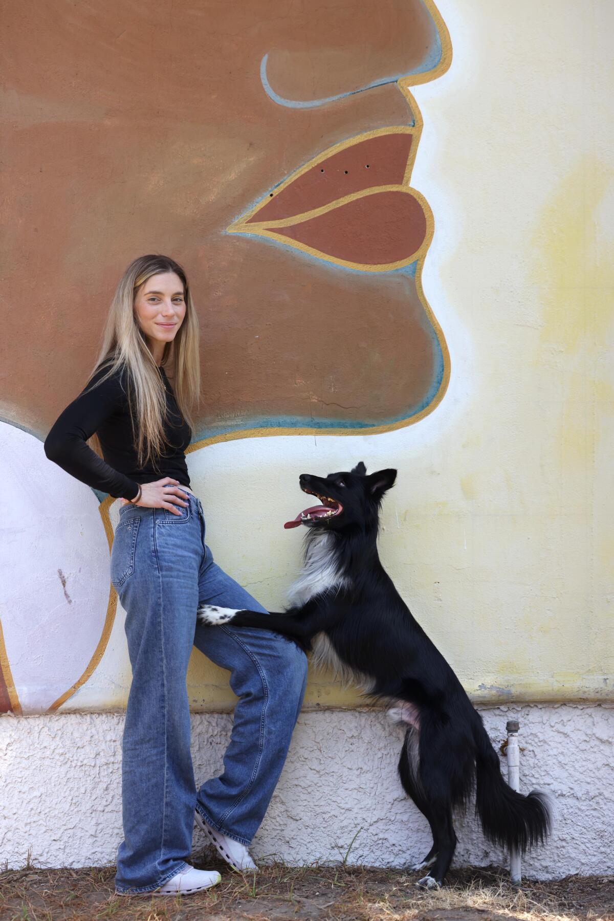 Woman poses with her dog leaning against her leg in front of a mural