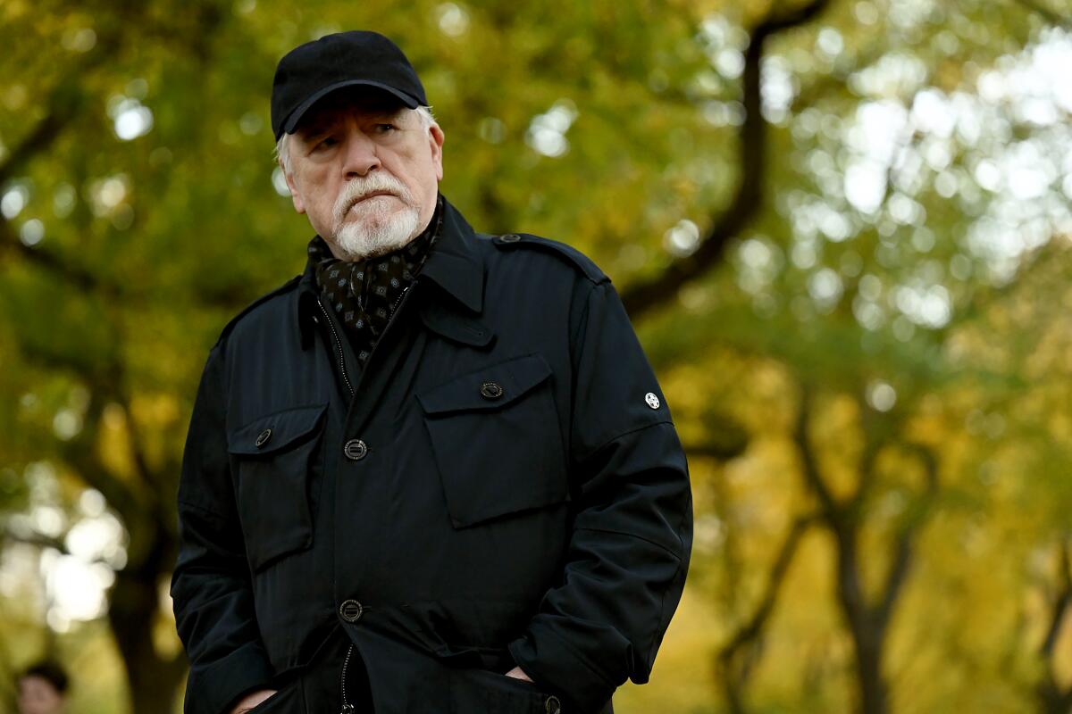 Brian Cox (as Logan Roy) frowns while wearing a black cap and jacket in the woods.