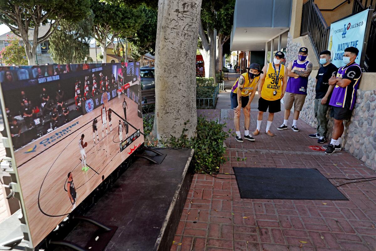 A group of people in Lakers jerseys watch a basketball game on an outdoor TV screen outside a bar