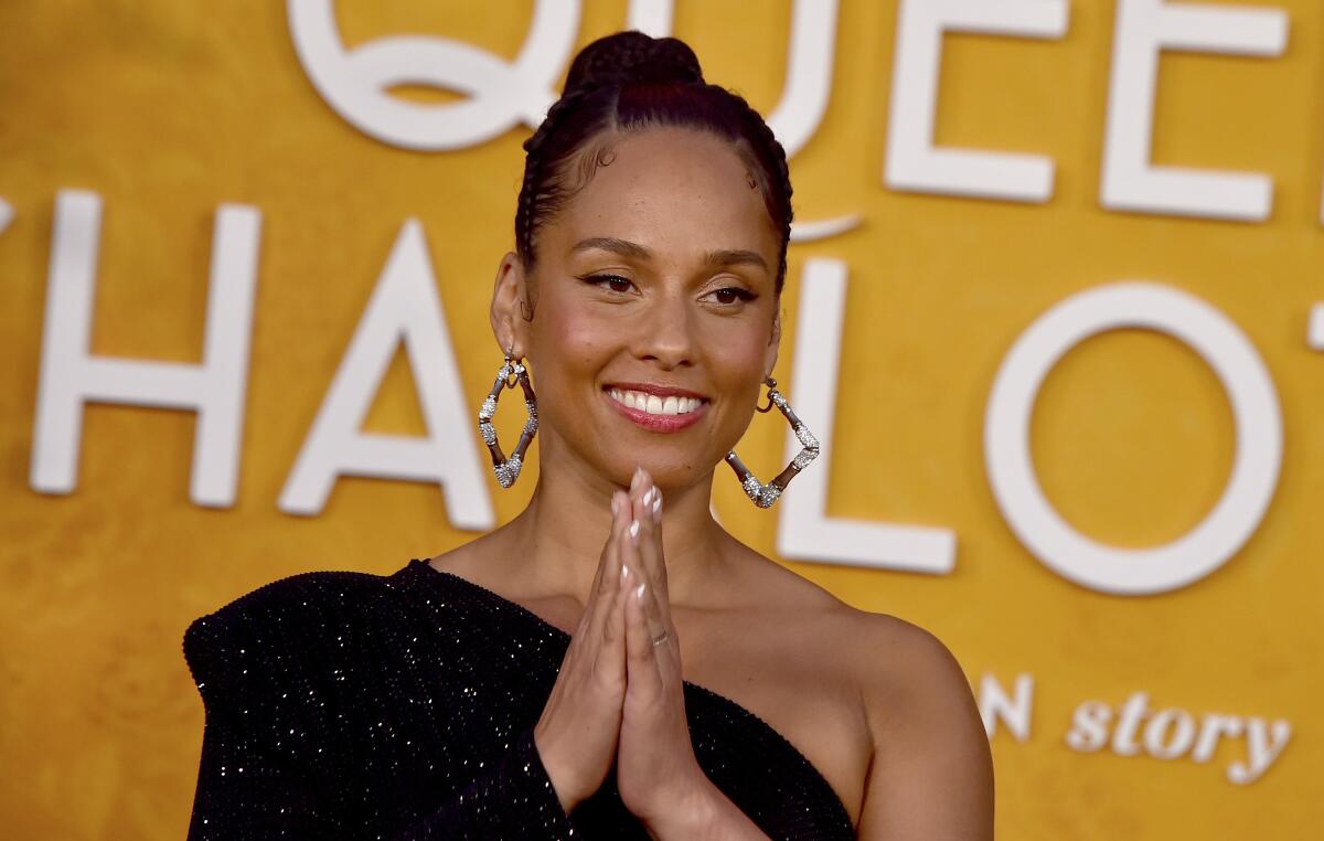 Alicia Keys, posing in a black off-the-shoulder dress, holds her hands in a prayer-like position.