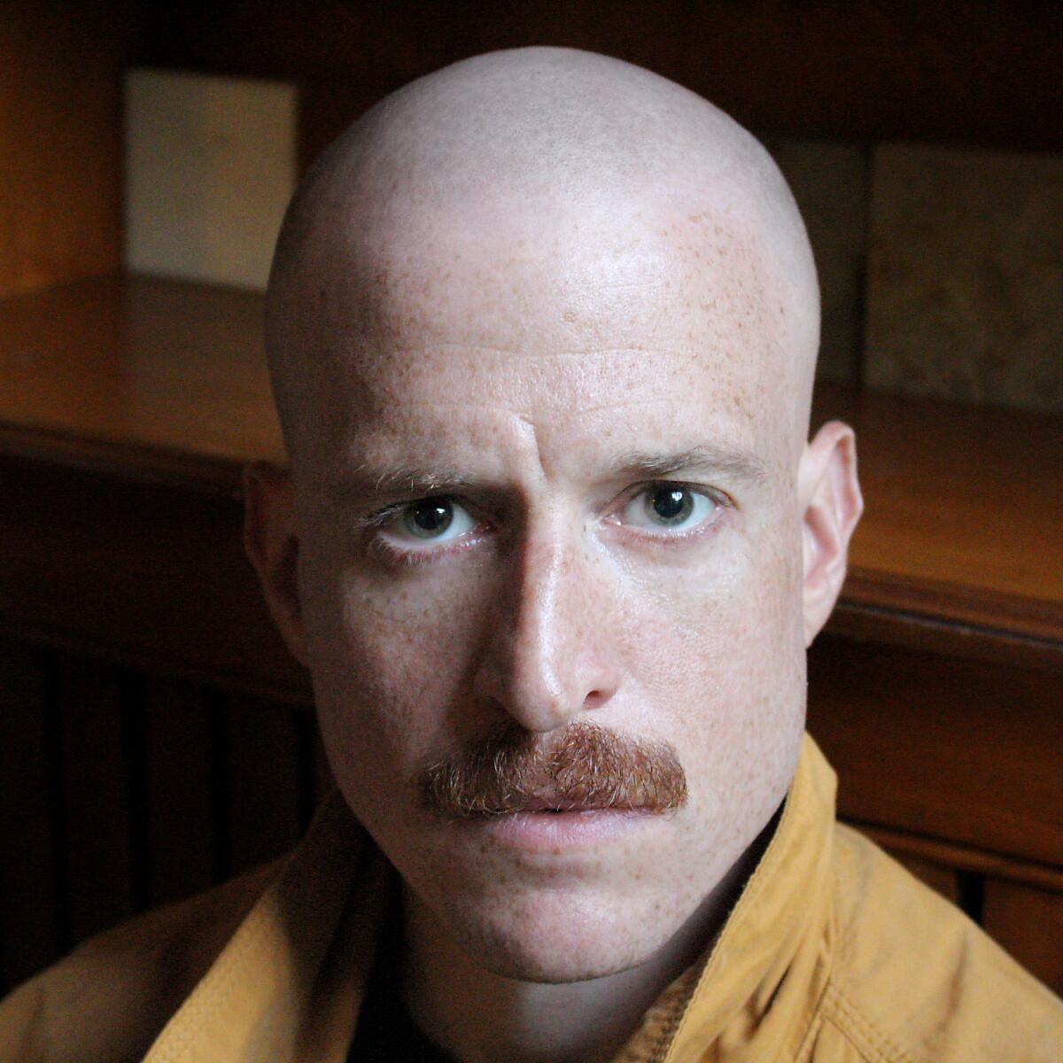 A man with a shaved head and red mustache in a yellow shirt, staring intensely.