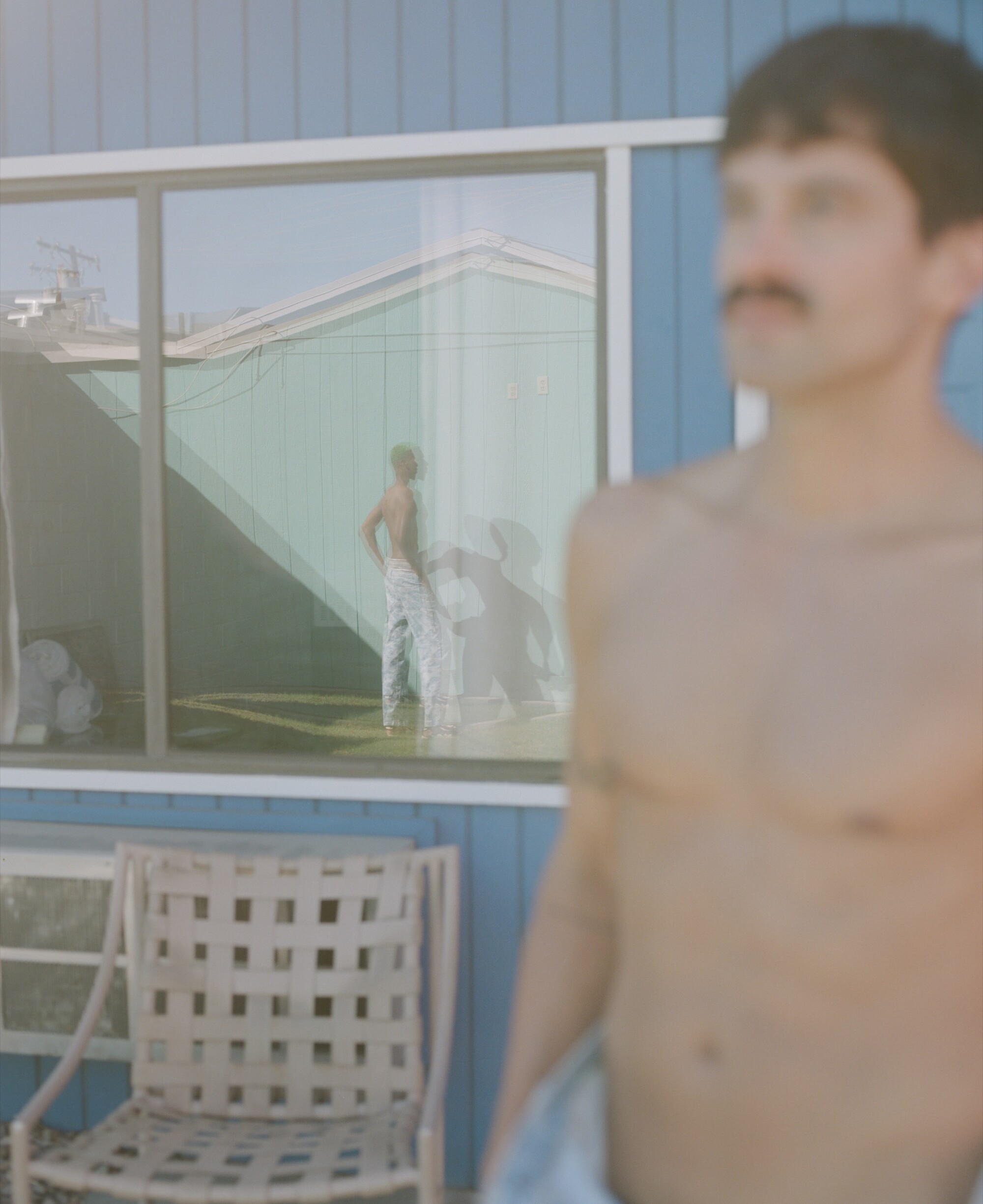 A mustached man stands looking into the distance at another man whose reflection is seen in a glass window.