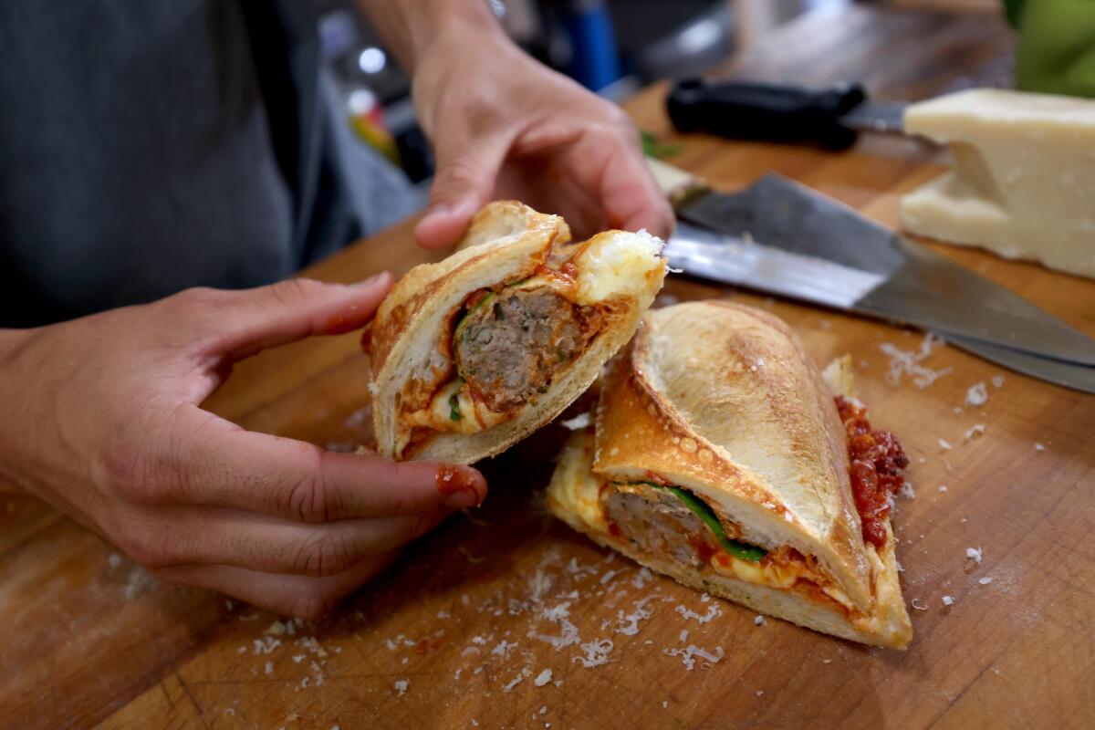 Owen Han holds one half of a completed meatball sandwich in his studio kitchen next to the other half on the cutting board.