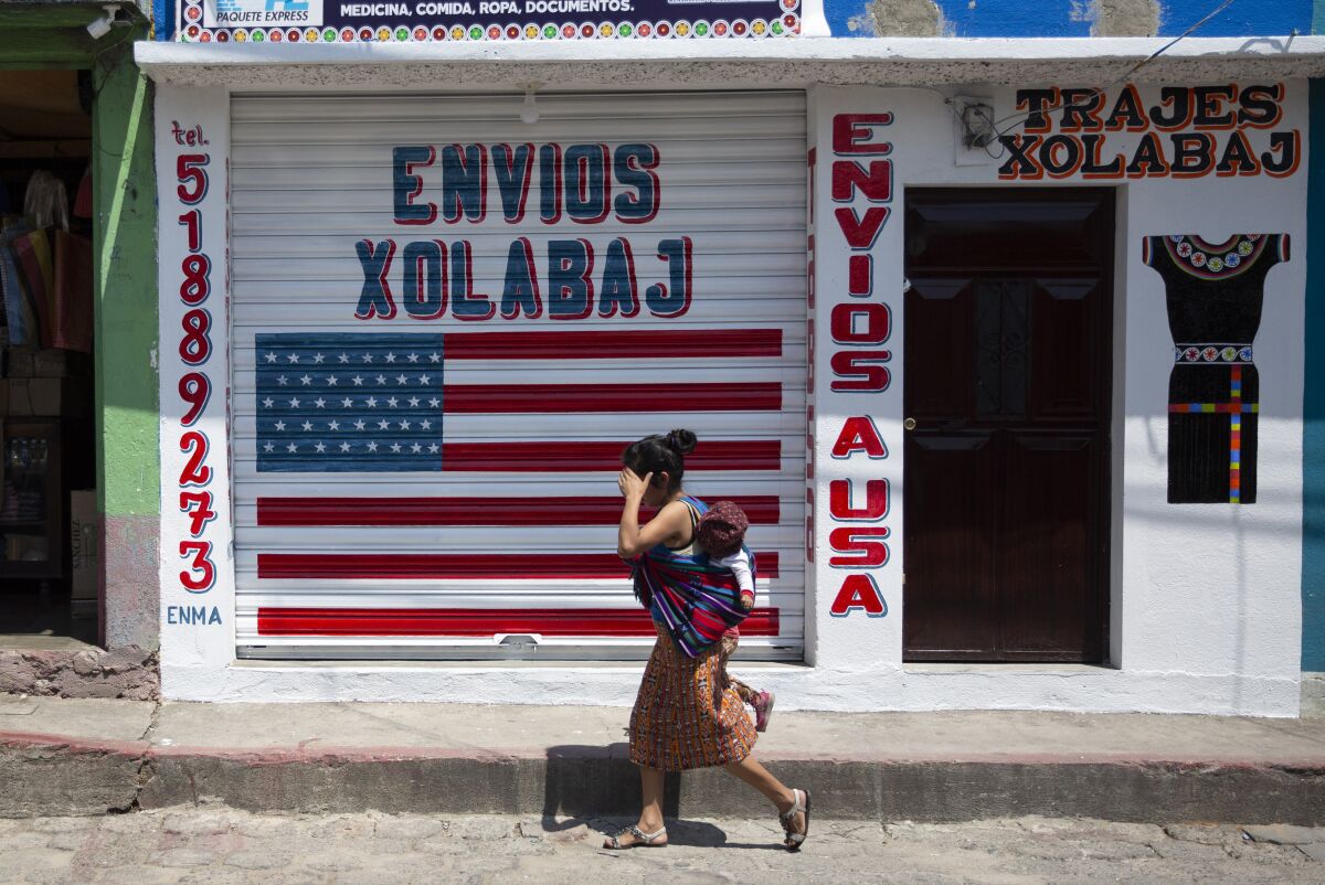 A woman carrying a child walks past a business labeled "Envios a USA"