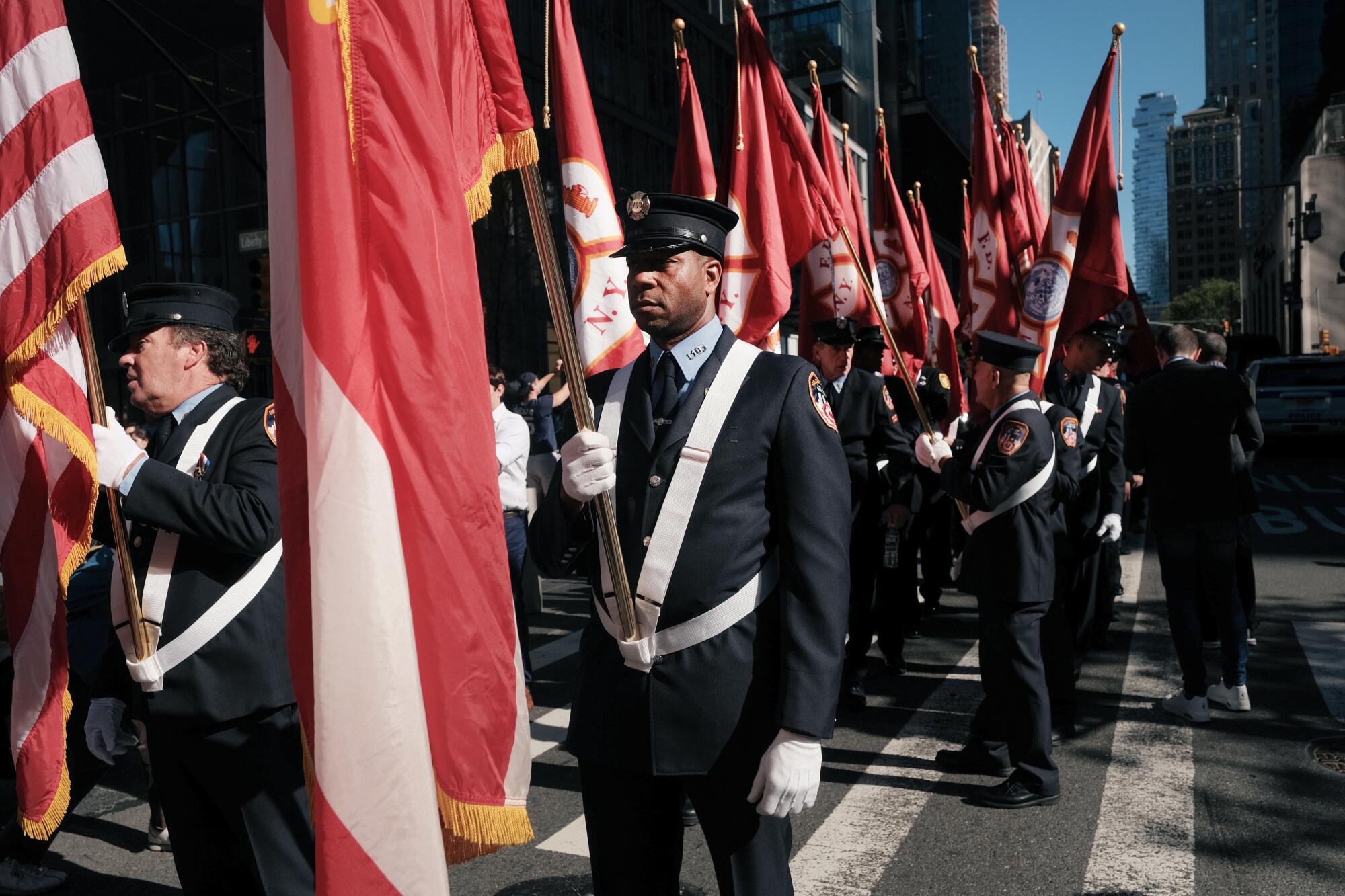 Firefighters in dress uniform gather in lower Manhattan carrying flags.