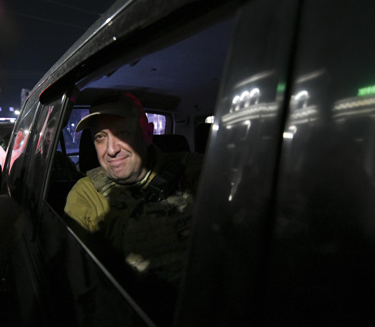 A man wearing a cap and olive-green clothing looks out the window of a vehicle 