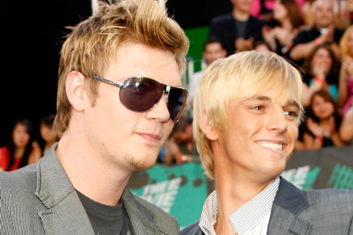 A blond man wearing sunglasses and posing with a younger blond man wearing a matching gray suit