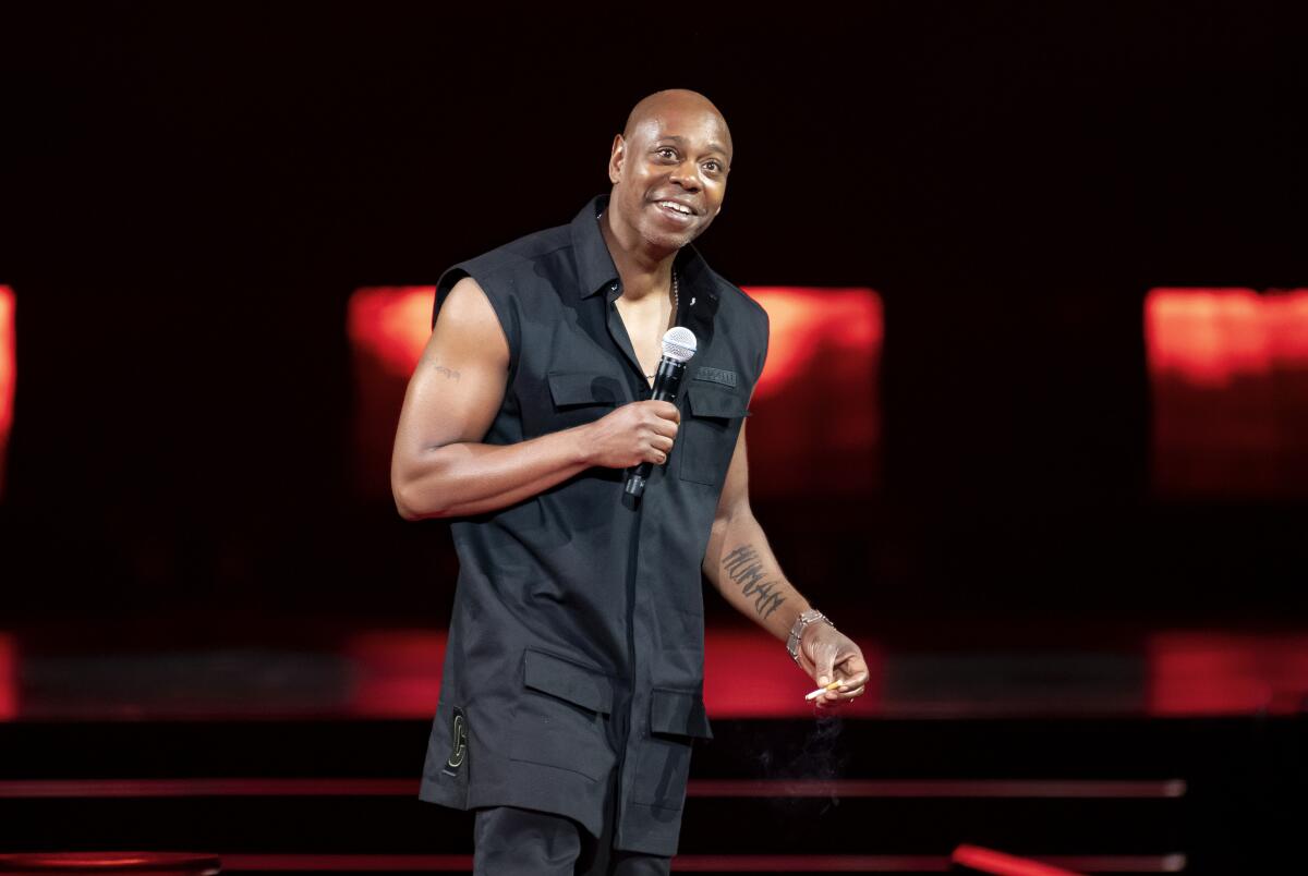 Comedian Dave Chappelle wearing a sleeveless jacket and holding a microphone onstage