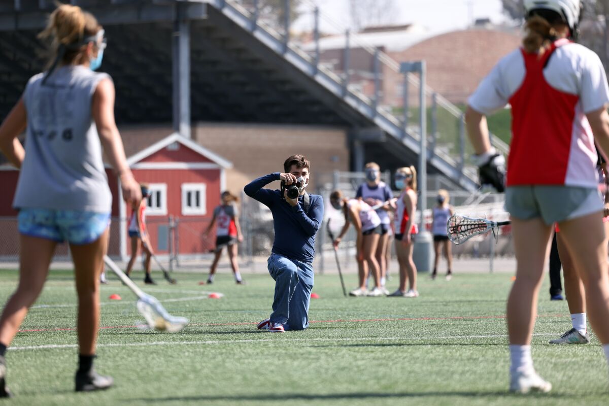 A photographer kneels on a lacrosse field while girls practice