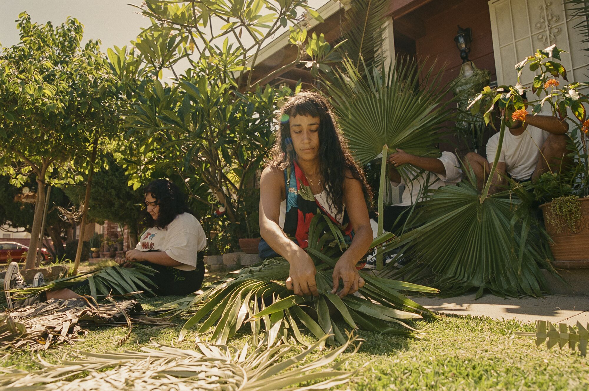 Several people sit on grass, cutting and weaving palms.