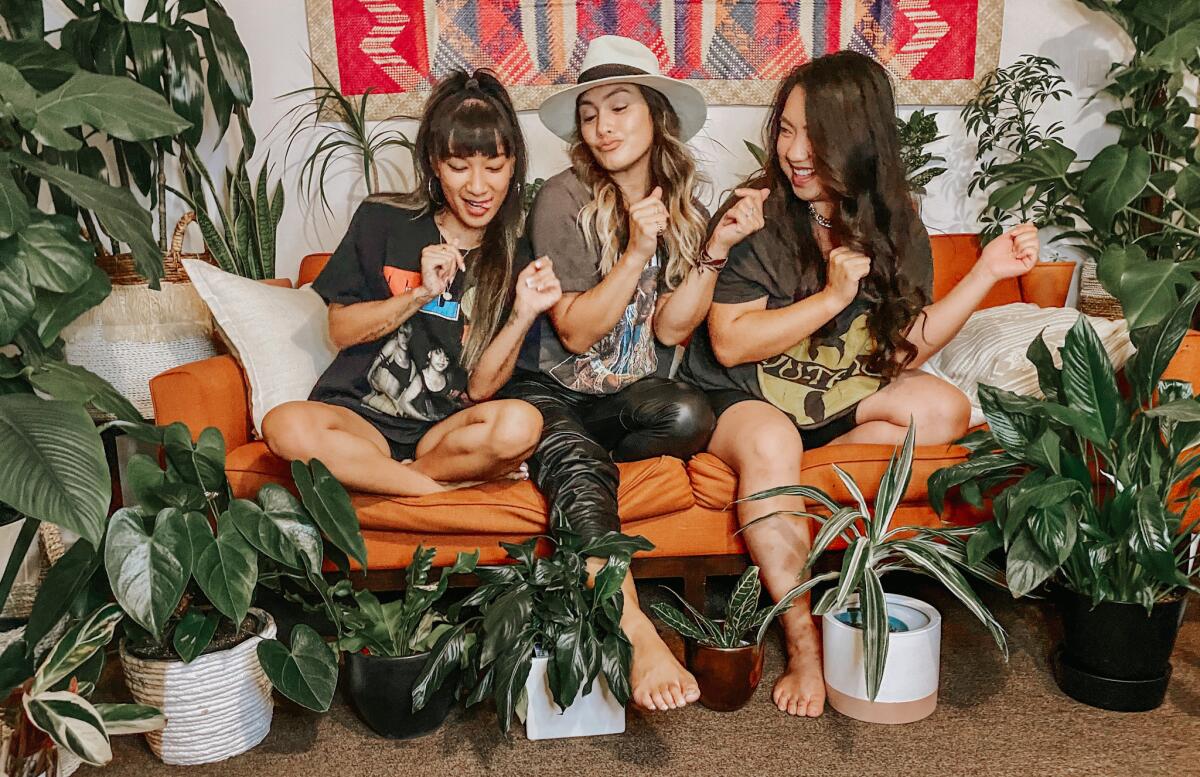 Three casually dressed women sitting on an orange sofa dancing and surrounded by houseplants.