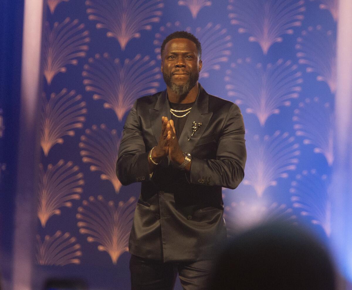 Kevin Hart puckers his lips and holds his hands together while wearing a shiny suit onstage.