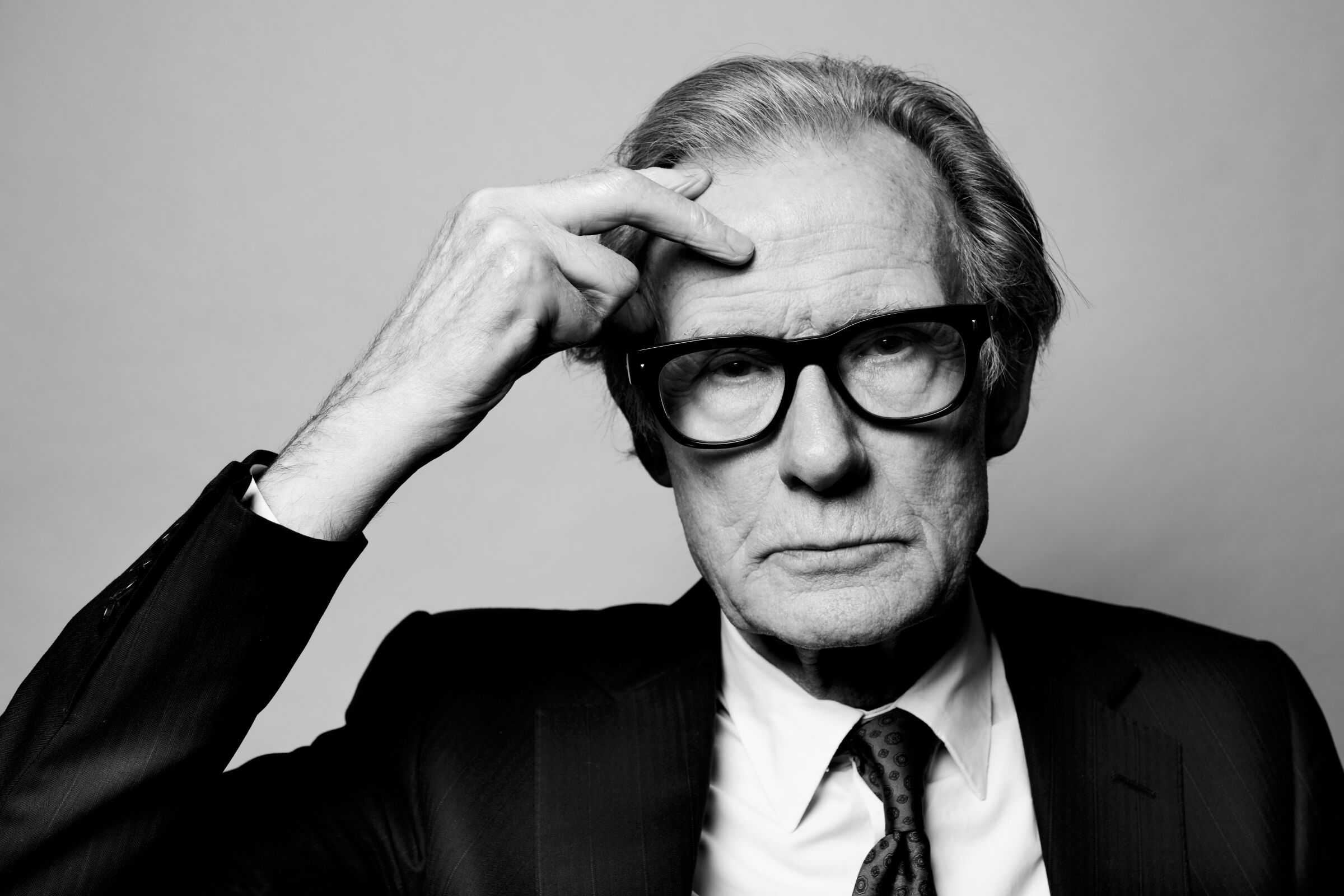 A black-and-white portrait of Bill Nighy, dressed in a dark suit and putting a hand to his forehead. 
