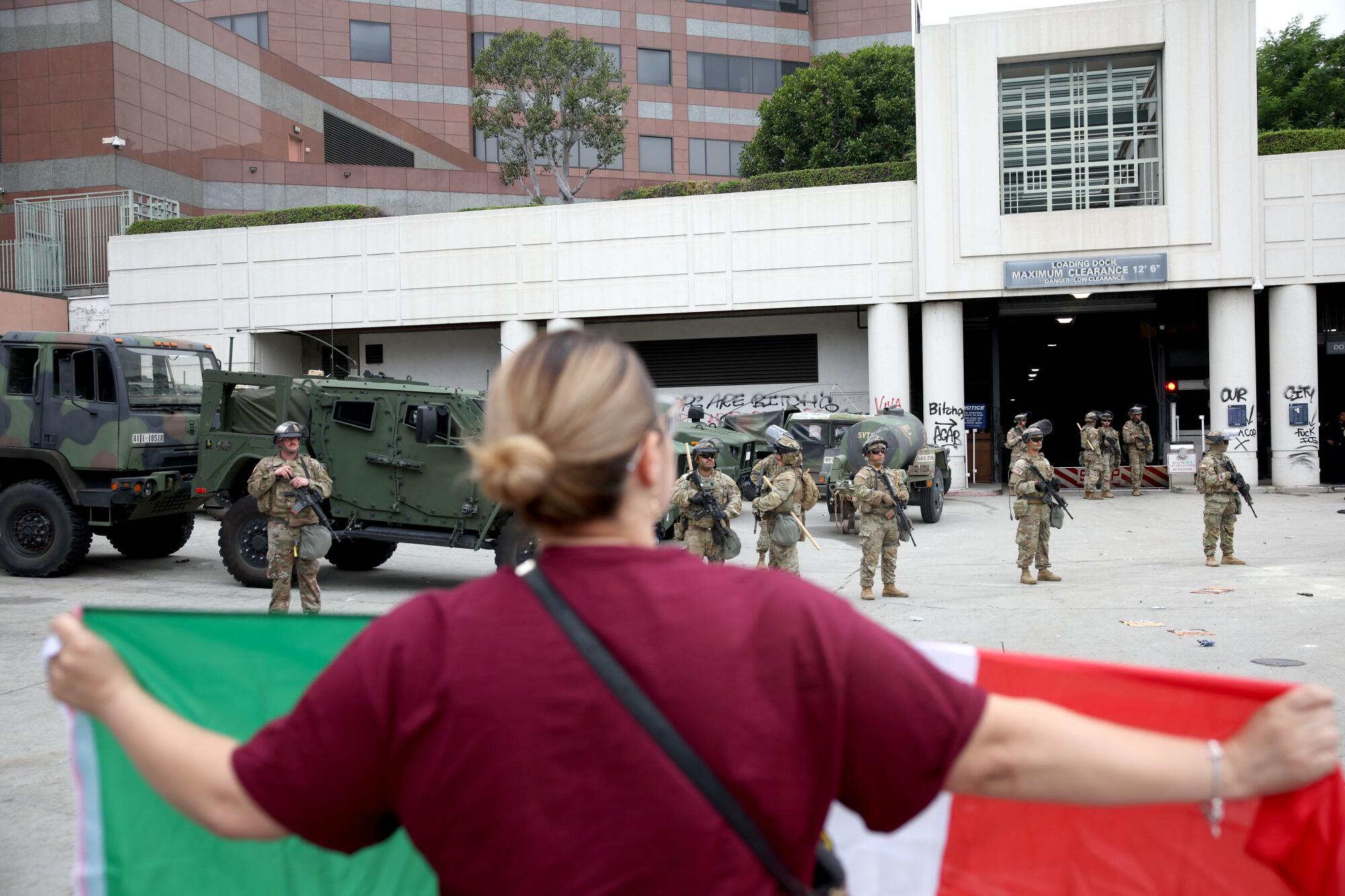A woman in a red shirt is shown from behind holding a green, white and red flag facing armed people in military uniform